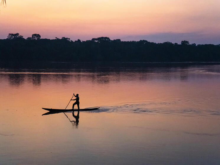 Someone standing up paddling on a slim canoe along a river at sunset with pink and purple hues