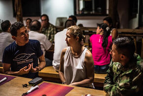 A woman sitting between two men at a dinner table in conversation with additional people behind them at other tables