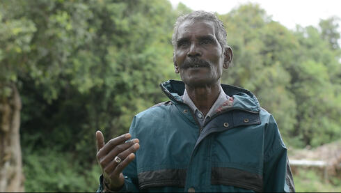 Portrait of a man in a teal jacket standing in front of a lush green forest